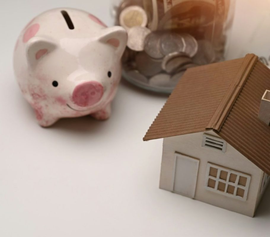 Sparschwein und ein Miniaturhaus neben einem Geldglas mit Münzen.