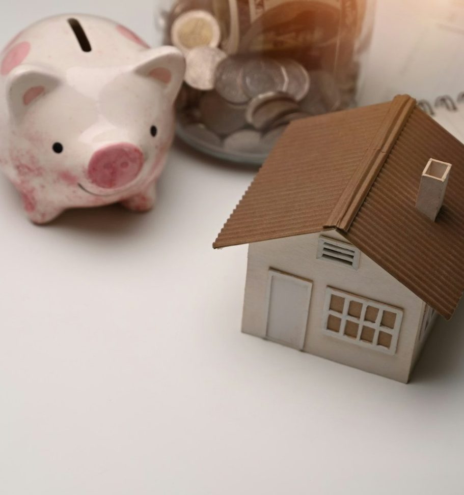 Sparschwein und ein Miniaturhaus neben einem Geldglas mit Münzen.